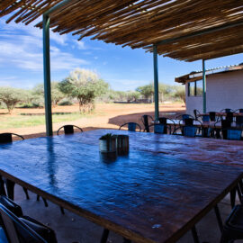 Large square wooden table with lots of chairs around it standing on the lodge porch with roof covering it
